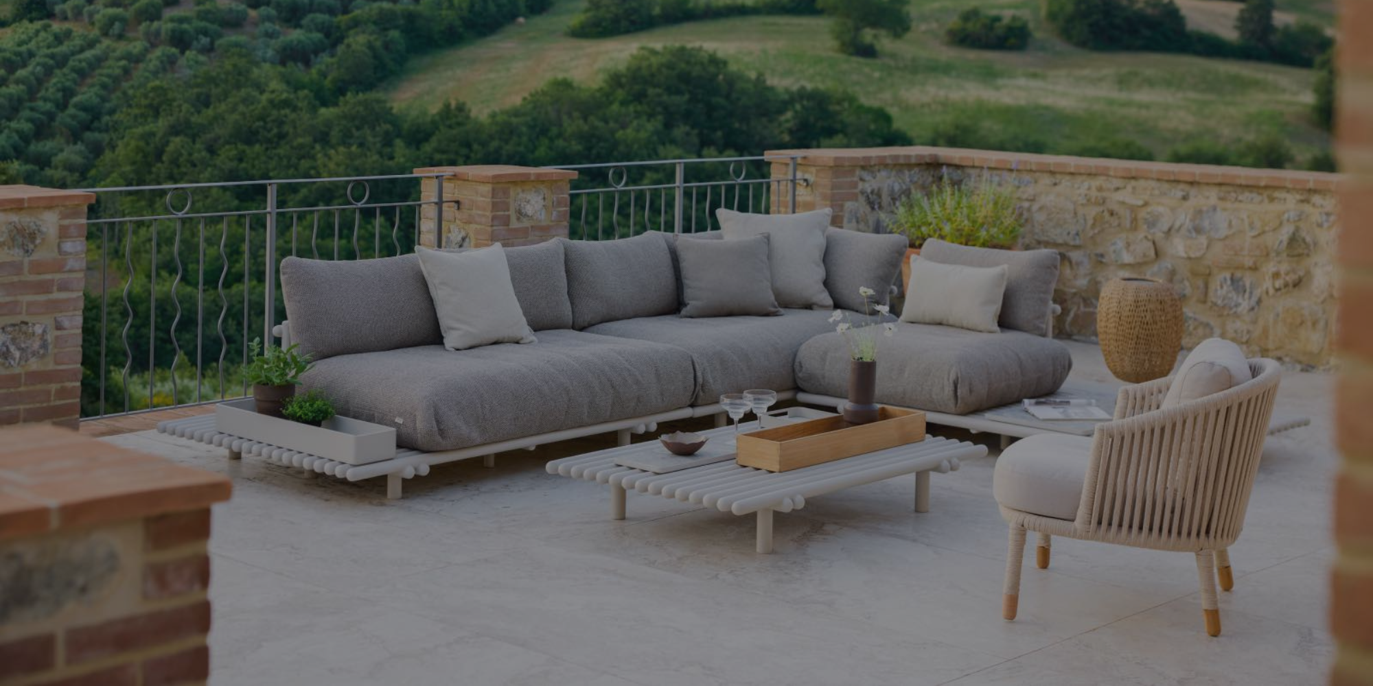 Outdoor patio with gray sectional sofa, coffee table, and chair on a stone patio with green landscape in the background.

